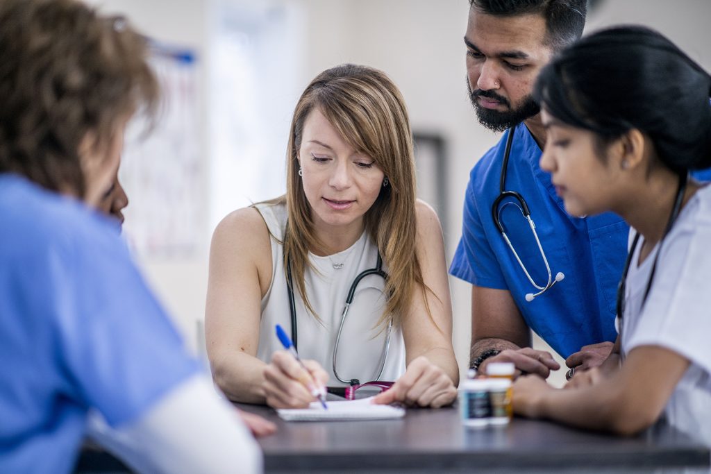 A multi-ethnic group of doctors are indoors in an office. They are gathered around a table for a meeting. One woman is taking notes.